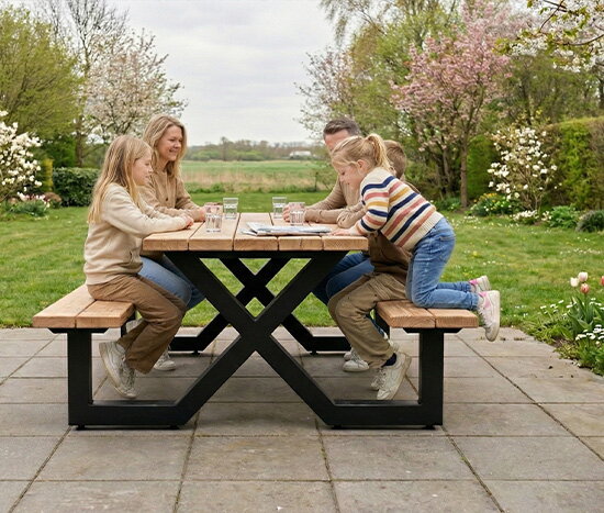 Une famille est assise autour d&rsquo;une table de pique-nique en bois sur une terrasse carrel&eacute;e, dans un jardin verdoyant et fleuri, tandis que deux enfants sont appuy&eacute;s &agrave; la table, en face de leurs parents.